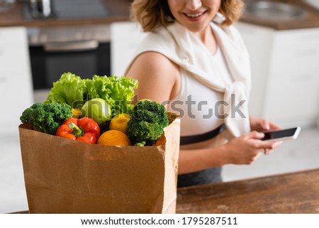 selective focus of paper bag with groceries near woman holding smartphone with blank screen