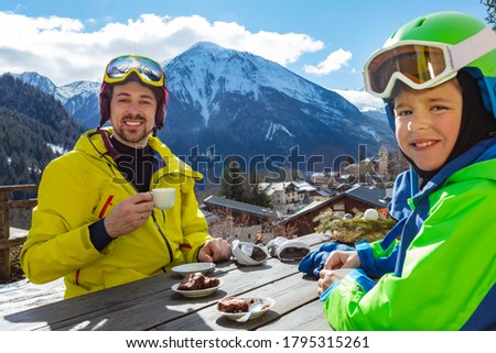 Portrait of a little boy in ski outfit sit and enjoy lunch break with father over mountain view after skiing