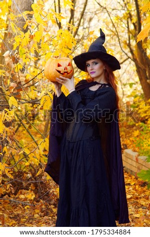 a girl in a black witch costume and pointed hat in an autumn yellow Park on Halloween with a pumpkin in her hands