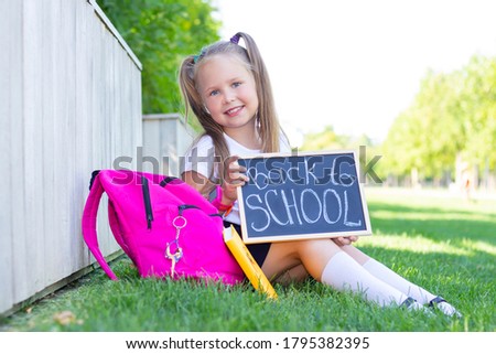 schoolgirl sits on the grass, school backpack. Holds a sign in his hands with the inscription back to school