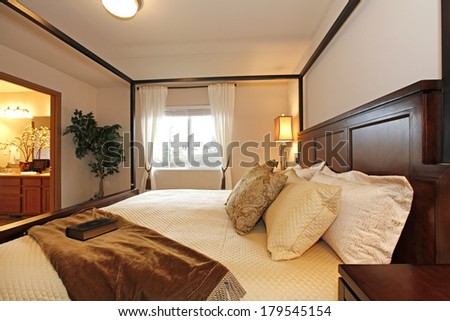 Light tones bedroom. Furnished with a queen size bed with high posts, nightstands. Decorated with a green tree. View of the bathroom