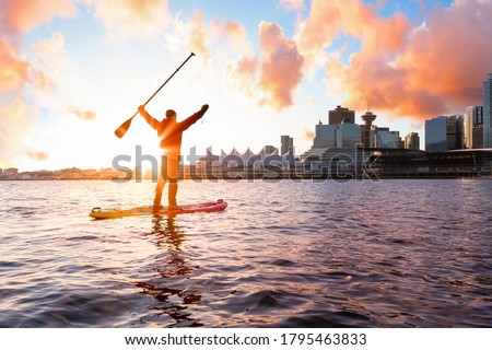Adventurous man is paddle boarding near Downtown City during a vibrant winter sunrise. Taken in Coal Harbour, Vancouver, British Columbia, Canada. Colorful Sky Overlay