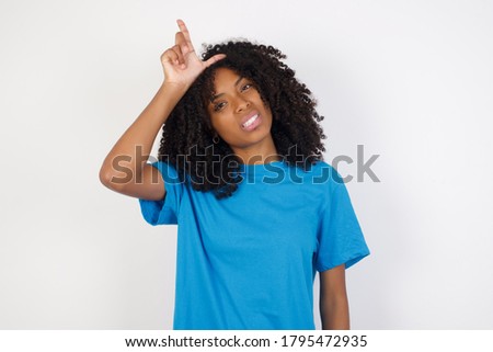 Young african woman with curly hair wearing casual blue shirt  making fun of people with fingers on forehead doing loser gesture mocking and insulting.