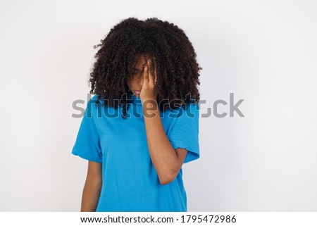 Tired overworked Young african woman with curly hair wearing casual blue shirt  has sleepy expression, gloomy look, covers face with hand, has eyes shut, gasps from tiredness, fatigue after party
