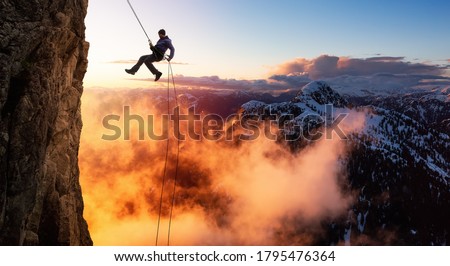 Epic Adventurous Extreme Sport Composite of Rock Climbing Man Rappelling from a Cliff. Mountain Landscape Background from British Columbia, Canada. Concept: Explore, Hike, Adventure, Lifestyle Royalty-Free Stock Photo #1795476364