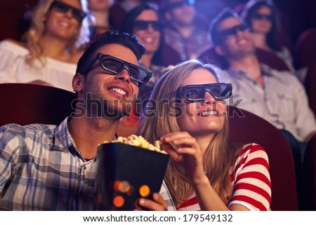 Happy couple sitting in movie theater, watching 3D movie, eating popcorn, smiling. Royalty-Free Stock Photo #179549132
