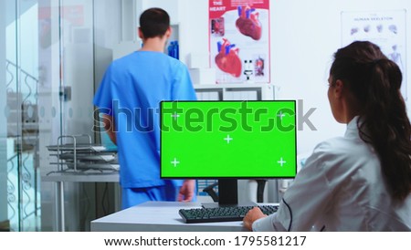 Physician using computer with chroma key and assistant holding patient x-ray in hospital cabinet. Desktop with replaceable green screen in medical clinic while doctor is checking patient radiography