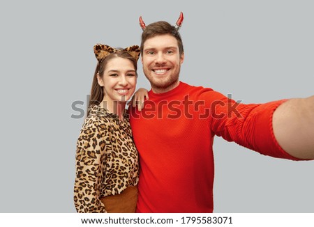 holiday and people concept - happy smiling couple in halloween costumes of devil and leopard taking selfie over grey background