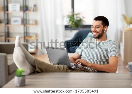 technology, remote job and lifestyle concept - happy smiling man with laptop computer and earphones resting feet on table at home office