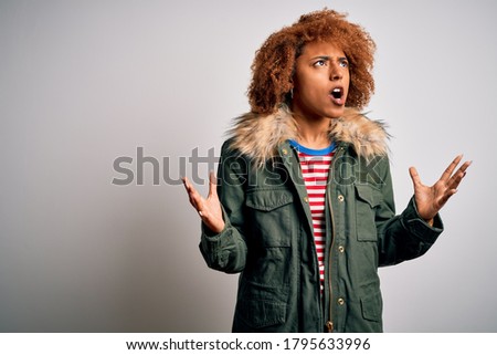 Young beautiful African American woman with curly hair wearing green casual winter coat crazy and mad shouting and yelling with aggressive expression and arms raised. Frustration concept.
