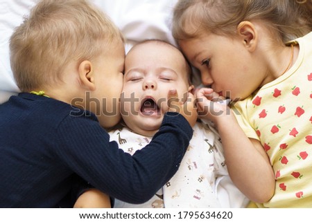 Little smiling kids playing together sitting on bed. Brother and sister kiss a newborn.Toddler kid meeting new born sibling. Infant sleeping in white bouncer under a blanket. Children with small age