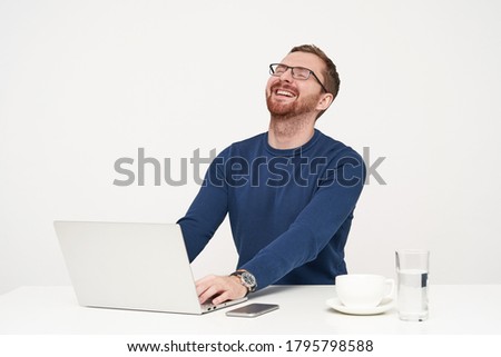 Joyful young bearded fair-haired man throwing back his head while laughing and keeping eyes closed, hearing funny joke while working with his laptop over white background