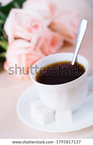 Cup of strong black tea stands on a saucer on a table. Pink roses in the background.