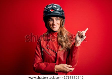 Young beautiful brunette motrocyclist woman wearing moto helmet over red background with a big smile on face, pointing with hand and finger to the side looking at the camera.