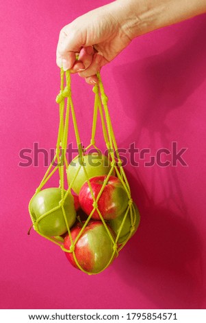 Hand holds a bag of string bag with ripe apples on a pink background. Eco-friendly packaging. Reusable. Copy space.