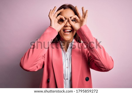 Young beautiful brunette woman wearing jacket standing over isolated pink background doing ok gesture like binoculars sticking tongue out, eyes looking through fingers. Crazy expression.