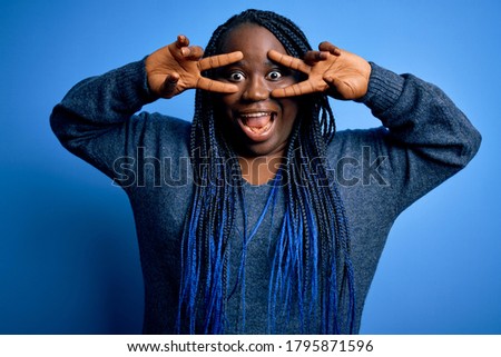 African american plus size woman with braids wearing casual sweater over blue background Doing peace symbol with fingers over face, smiling cheerful showing victory