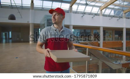 Young delivery guy carrying pizza box in empty office hallway. Courier in uniform holding meal box delivering order in shopping mall or business center