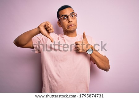 Handsome african american man wearing casual t-shirt and glasses over pink background Doing thumbs up and down, disagreement and agreement expression. Crazy conflict