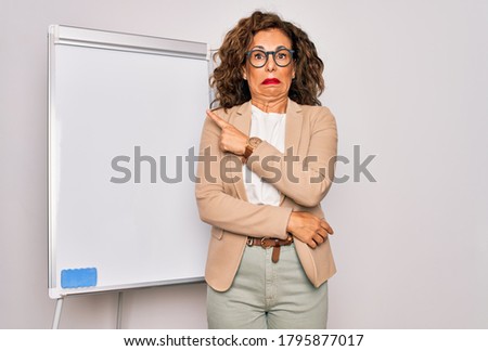 Middle age senior business woman standing on seminar presentation by magnectic blackboard Pointing aside worried and nervous with forefinger, concerned and surprised expression