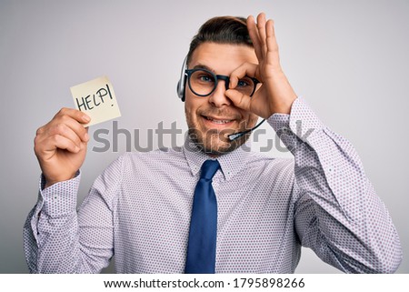 Young call center operator business man with blue eyes offering support holding paper note with happy face smiling doing ok sign with hand on eye looking through fingers