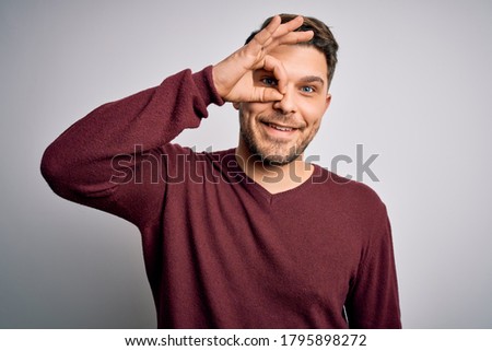 Young man with blue eyes wearing casual sweater standing over isolated background doing ok gesture with hand smiling, eye looking through fingers with happy face.