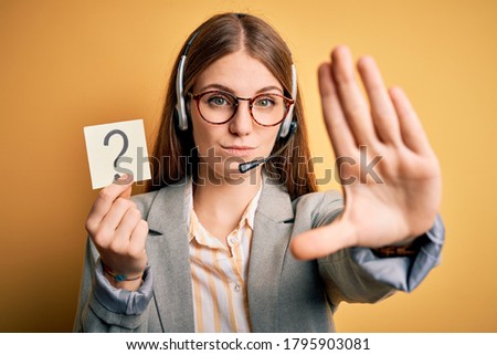 Young redhead call center agent woman using headset holding question mark reminder with open hand doing stop sign with serious and confident expression, defense gesture