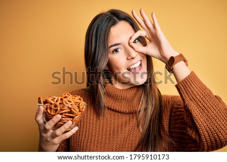 Young beautiful girl eating baked german pretzel standing over isolated yellow background with happy face smiling doing ok sign with hand on eye looking through fingers