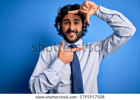 Young handsome call center agent man with beard working using headset over blue background smiling making frame with hands and fingers with happy face. Creativity and photography concept.