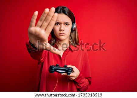Young beautiful brunette gamer woman playing video game using joystick and headphones with open hand doing stop sign with serious and confident expression, defense gesture
