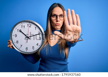 Young blonde woman with blue eyes holding big minute clock over isolated background with open hand doing stop sign with serious and confident expression, defense gesture
