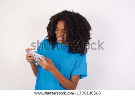 Young african woman with curly hair wearing casual blue shirt standing against white  background holding in hands cell playing video games or chatting 