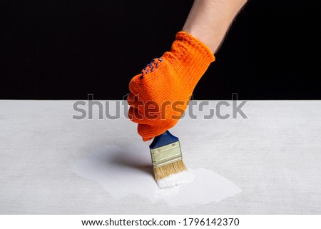 painting with white paint with a brush with a blue handle which is in hand in an orange glove on a black background