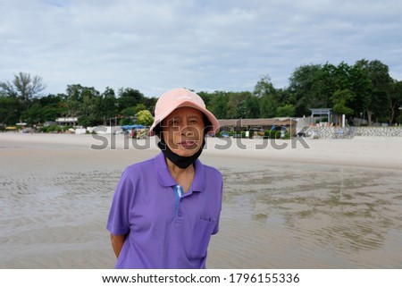 An old asian lady posing for a picture with a cloth face mask under her chin on the pristine Hua Hin beach, Thailand.