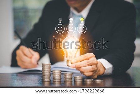 Businessman hand holding light bulb with Emoticon face icon and Coins stack on the wooden table , Saving ideas and investment budget, Creative ideas concept of saving money  concept, Copy space