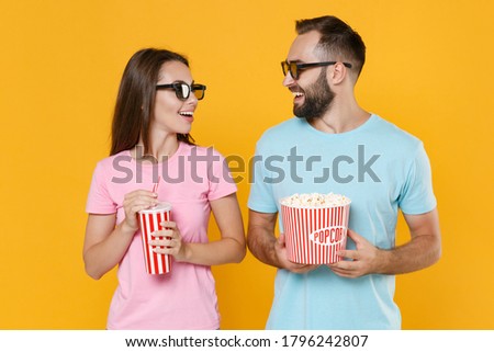 Cheerful couple two friends guy girl in t-shirts 3d glasses isolated on yellow background. People in cinema, lifestyle concept. Watching movie film hold bucket popcorn cup soda looking at each other.