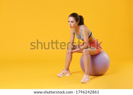 Full length portrait of strong young fitness sporty woman in sportswear working out isolated on yellow background studio. Workout sport motivation lifestyle concept. Sit on fit ball looking aside