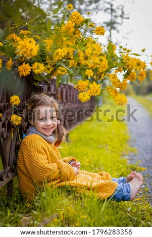 A smiling little girl sitting near the fence under the yellow big flowers