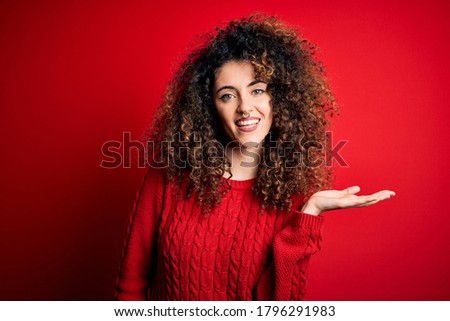 Young beautiful woman with curly hair and piercing wearing casual red sweater smiling cheerful presenting and pointing with palm of hand looking at the camera.