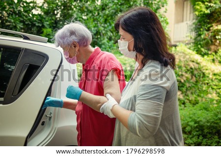 An attentive caregiver helps her elderly client to get into a car to travel for regular medical appointments. Both wear protective masks. Royalty-Free Stock Photo #1796298598