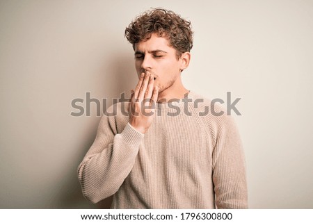 Young blond handsome man with curly hair wearing casual sweater over white background bored yawning tired covering mouth with hand. Restless and sleepiness.