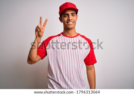 Young handsome african american sportsman wearing striped baseball t-shirt and cap smiling looking to the camera showing fingers doing victory sign. Number two.