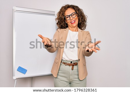Middle age senior business woman standing on seminar presentation by magnectic blackboard smiling cheerful offering hands giving assistance and acceptance.