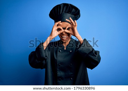 Young african american chef woman wearing cooker uniform and hat over blue background doing ok gesture like binoculars sticking tongue out, eyes looking through fingers. Crazy expression.