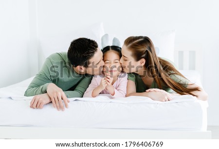 Enjoying free time together. Portrait of happy Asian family lying on white bed and kissing little daughter. Family holiday and togetherness, Love emotion.