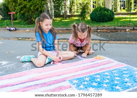 Two cute friends girls drawing American flag with colored chalks on the sidewalk near the house on sunny summer day. Kids painting outside. Creative development of children. Patriotic day concept