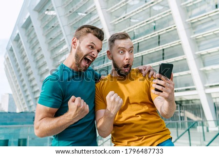 Two young bearded friends showing sincere emotions of joy about victory in internet lottery. Men celebrating money win in online sport gambling application with football stadium on the background. 