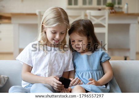 Happy children sitting on couch at living room watching clips or cartoons on mobile phone together, smiling elder girl showing cute little sister preschooler fun video on smartphone screen online