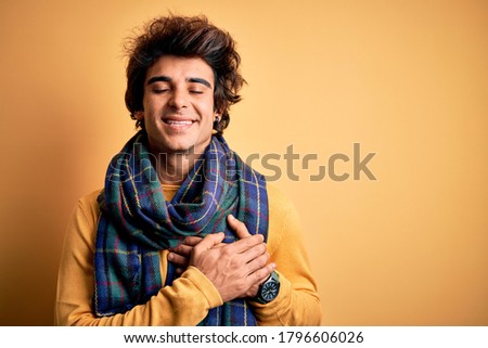 Young handsome man wearing casual scarf standing over isolated yellow background smiling with hands on chest with closed eyes and grateful gesture on face. Health concept.