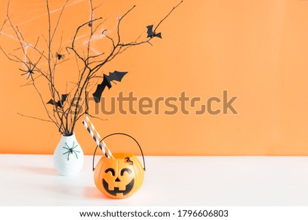 Halloween decorations, paper bats flying on tree branch and pumpkin bucket on table over orange background. Halloween background.